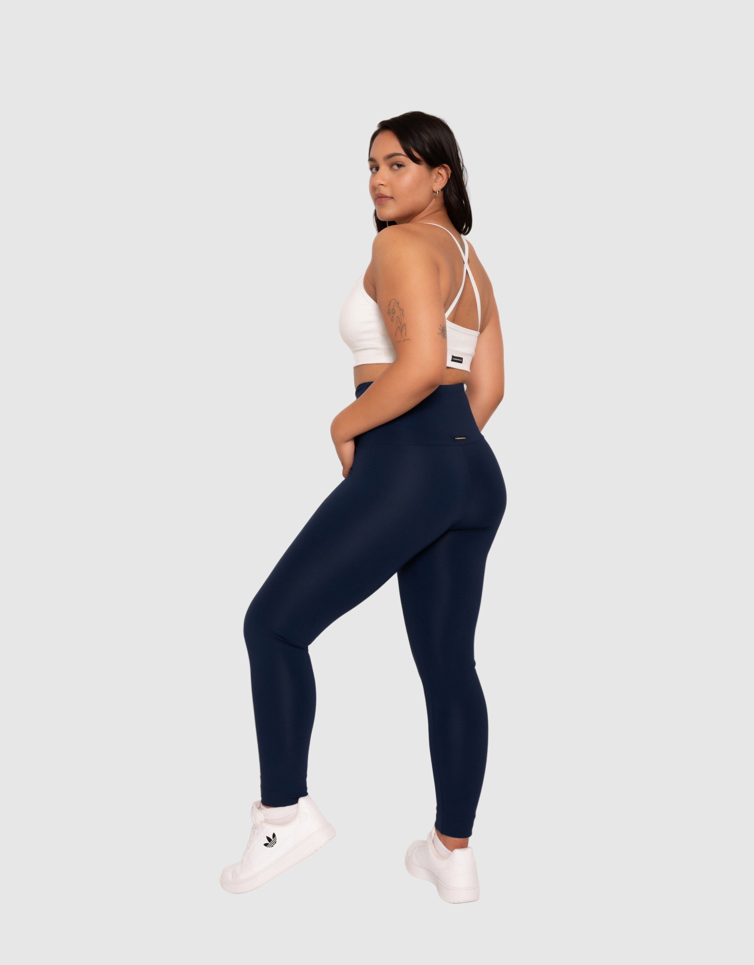 Woman wearing navy leggings and a white sports bra on a plain background